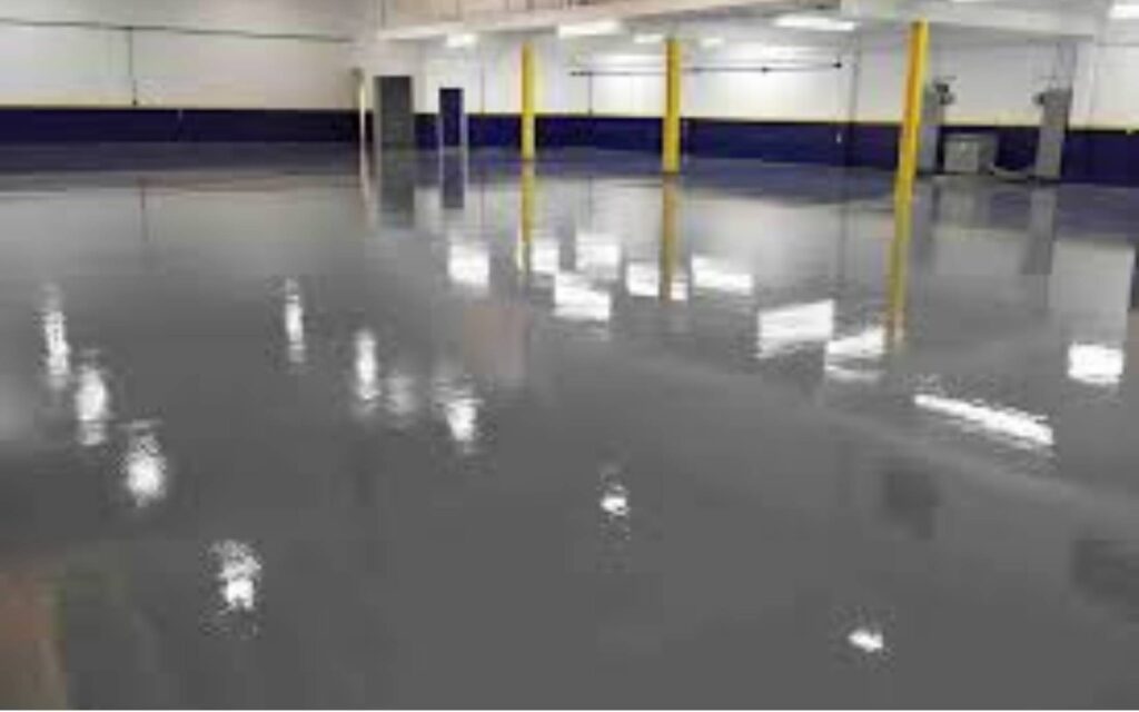 Spacious, empty warehouse with a glossy, reflective gray floor. Yellow pillars and dark blue lower walls contrast against white upper walls. Bright lighting.