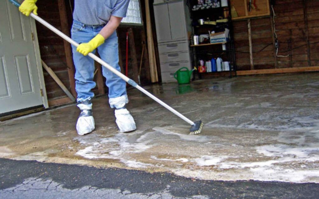 A person scrubbing a garage floor with a brush on a long handle. They are wearing yellow gloves and protective coverings on their shoes. Soap suds are visible.