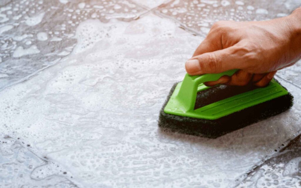 A hand scrubs soapy tiled floor with a green-handled sponge. The image conveys cleanliness and domestic care. Bubbles cover the gray tiles.