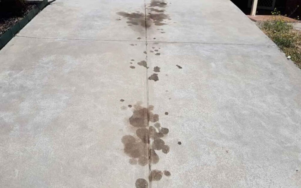 Concrete path with scattered dark oil stains running along the center. Flanked by grass on the right, creating a neglected and messy appearance.